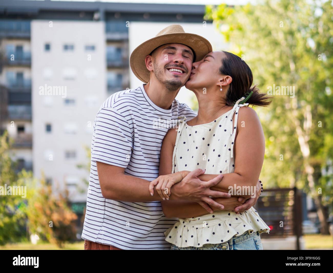 Fröhlicher Mann und Frau teilen eine herzliche Umarmung und einen Kuss, umgeben von Grün in einem Stadtpark. Stockfoto