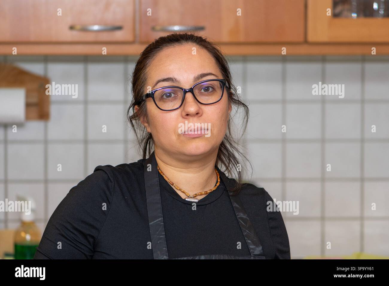 Eine Frau mit Brille und dunkler Schürze blickt in einer Kücheninterieur in die Kamera. Stockfoto