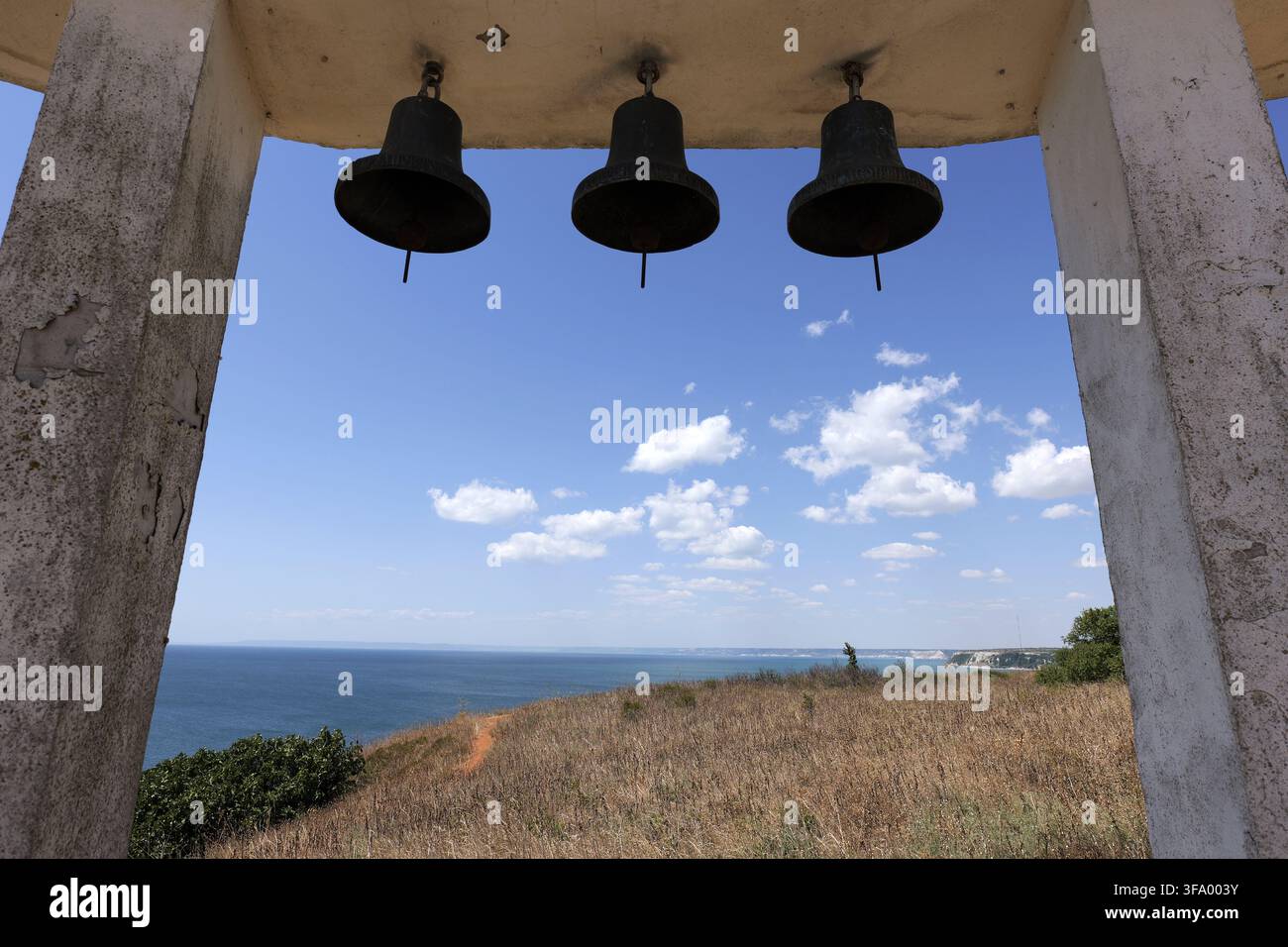 Drei Metallglocken, die in einem Beton-Glockenturm hängen, umrahmen einen Panoramablick auf das weite blaue Meer und die ferne Küstenlinie unter einem hellen Himmel mit Streuung Stockfoto