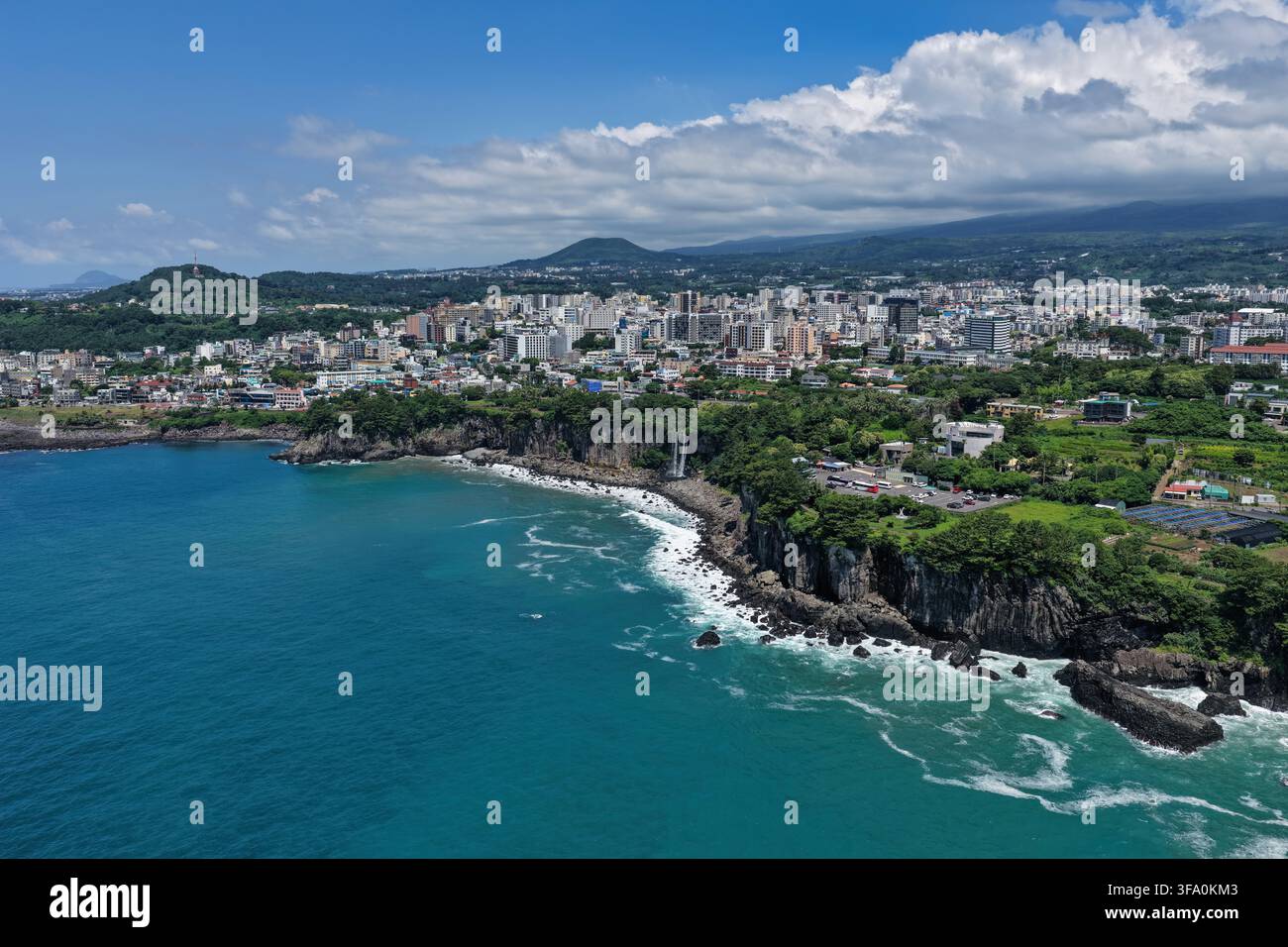 Luftaufnahme des Jeongbang-Wasserfalls, der über Küstenklippen ins Meer stürzt, mit der Stadt Seogwipo und üppiger Vegetation im Hintergrund in Südkorea Stockfoto