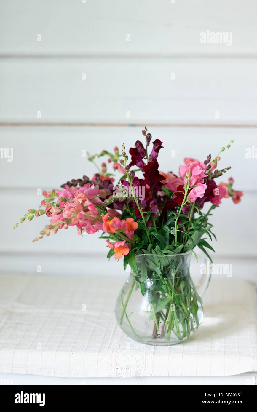 Buntes Bouquet aus rosa, korallenfarbenen und tief burgunderroten Blumen, arrangiert in einer klaren Glasvase auf einem weißen Tischtuch, vor einem weich gemalten Holz Stockfoto