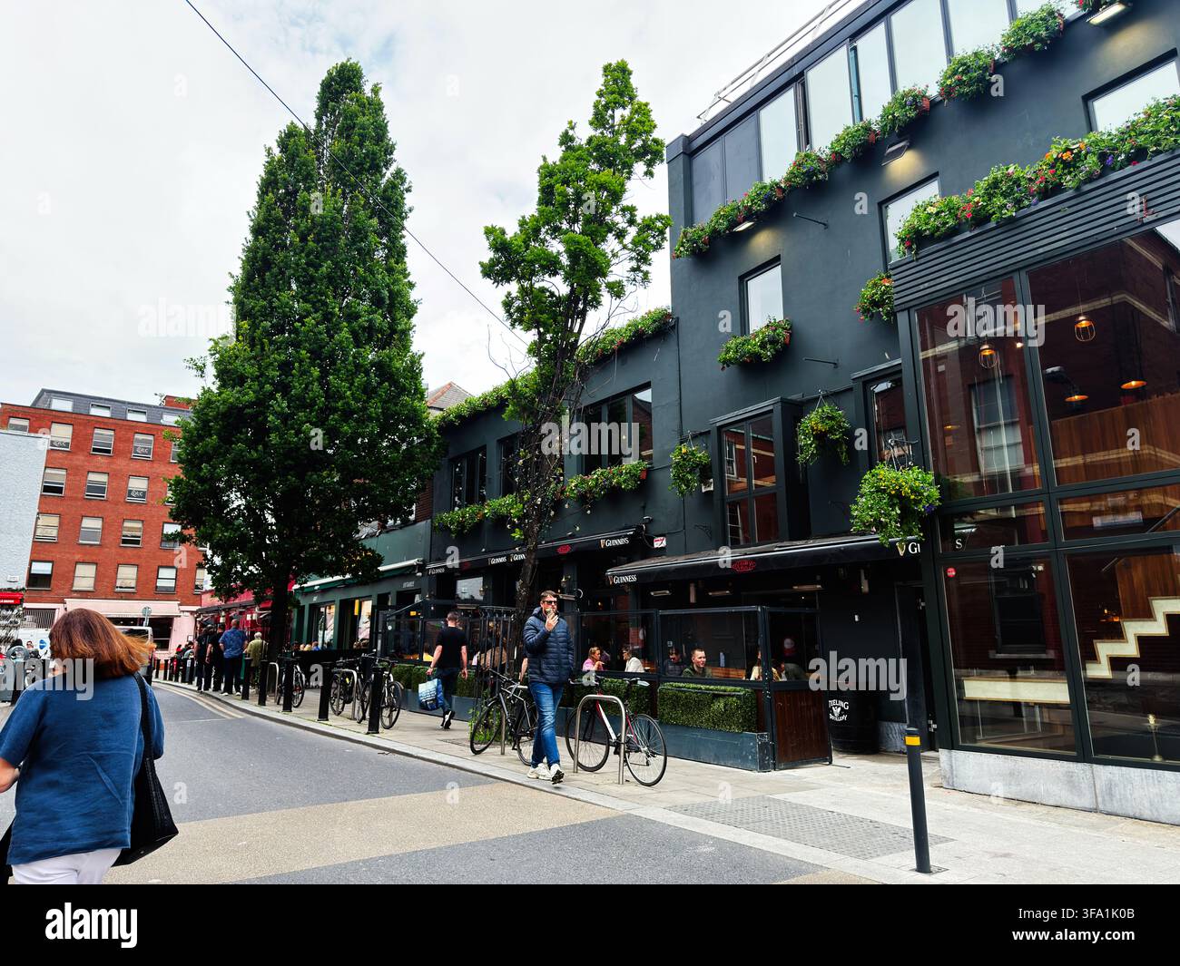 Dublin, Irland – 22. Mai 2026: Menschenmenge, die vor einem beliebten Dubliner Pub mit Pflanzen und Fahrrädern in der Nähe läuft und sitzt. Stockfoto