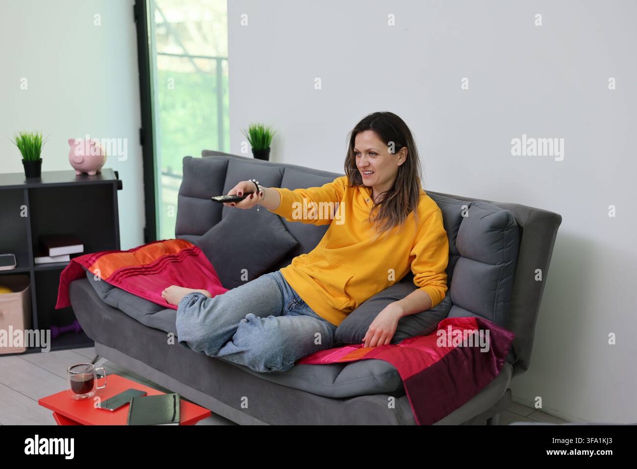Entspannte Frau in gelbem Sweatshirt, die auf grauem Sofa liegt und mit der Fernbedienung auf den Fernseher in einem hellen Wohnzimmer mit sanftem Tageslicht zeigt, farbenfroh Stockfoto