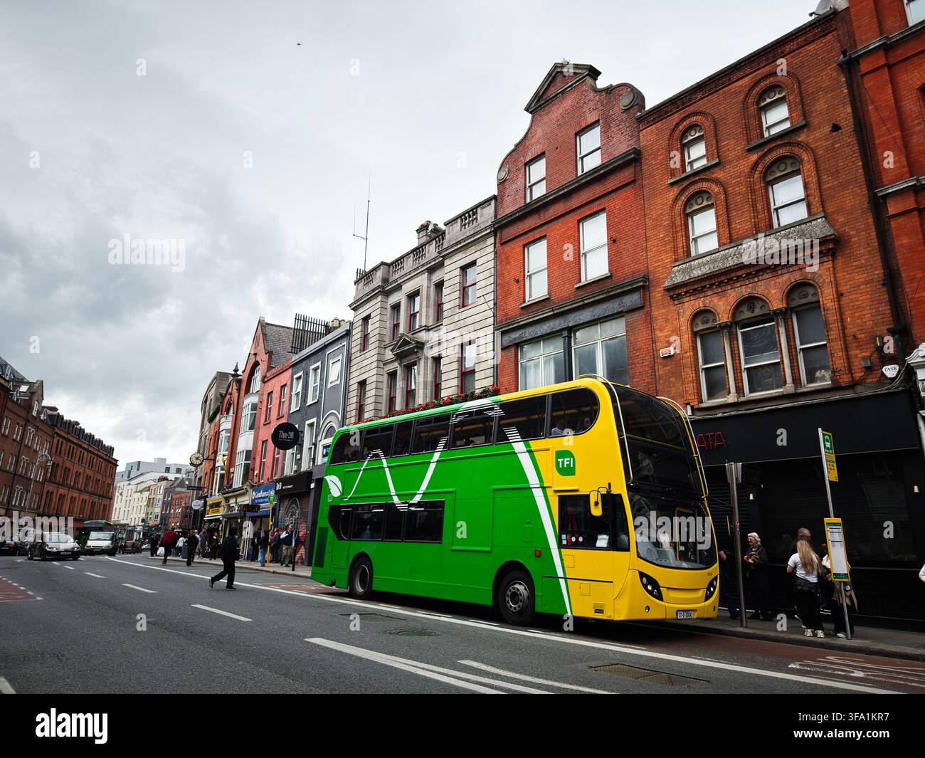 Dublin, Irland – 22. Mai 2026: Ein grüner und gelber Doppeldeckerbus steht auf einer Dubliner Straße, die von historischen Backsteinläden und Fußgängern gesäumt ist. Stockfoto