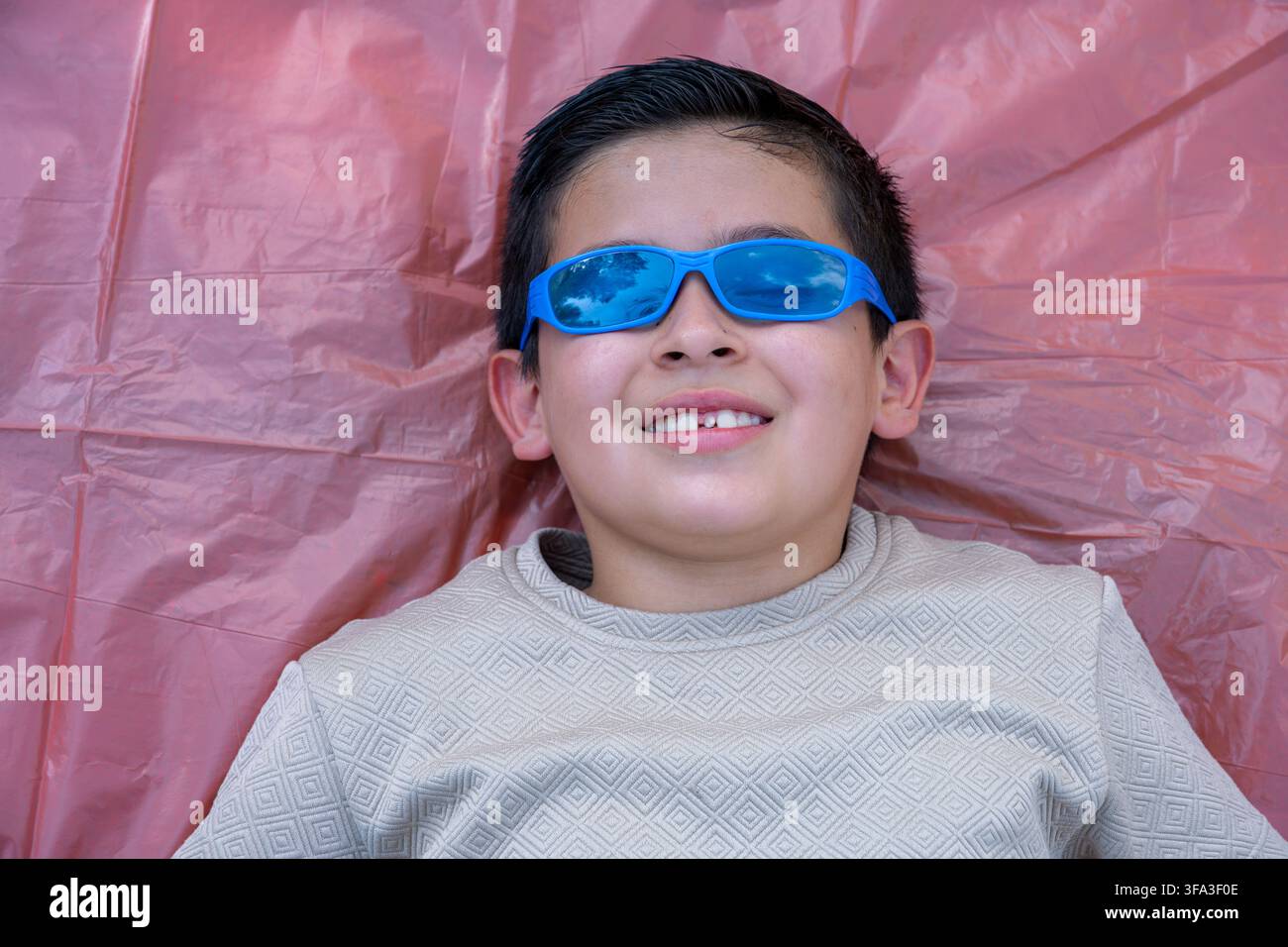 Junge mit blauer Sonnenbrille lächelt, liegt auf einem rosa Hintergrund, reflektiert den Himmel und die Wolken in seinen Gläsern, erlebt Freude und Entspannung während Stockfoto