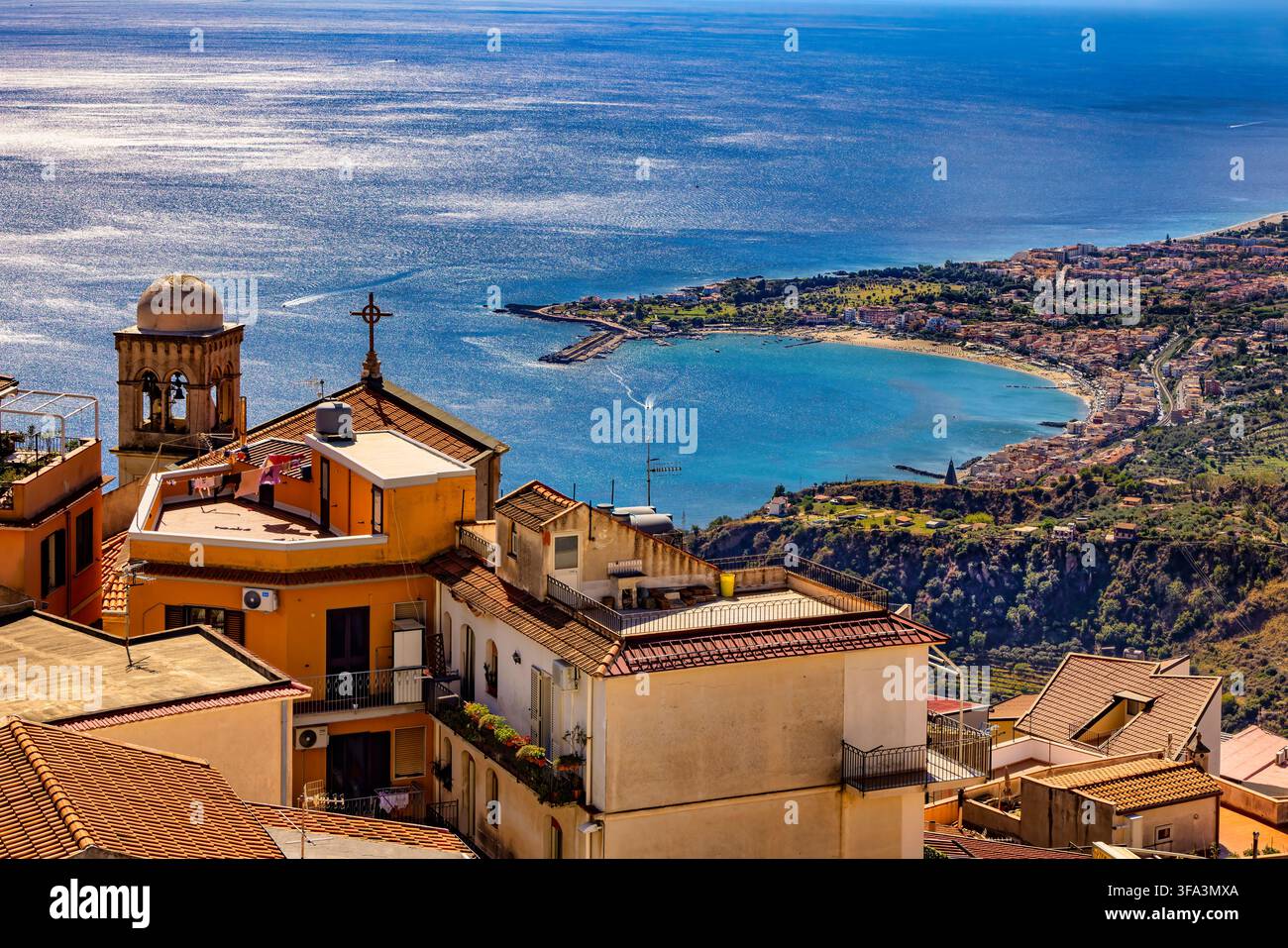 Giardini Naxos und die sizilianische Küste aus Taormina gesehen. Ein erhöhter Panoramablick führt über terracottafarbene Dächer und eine Kirche in Richtung der weiten Stockfoto