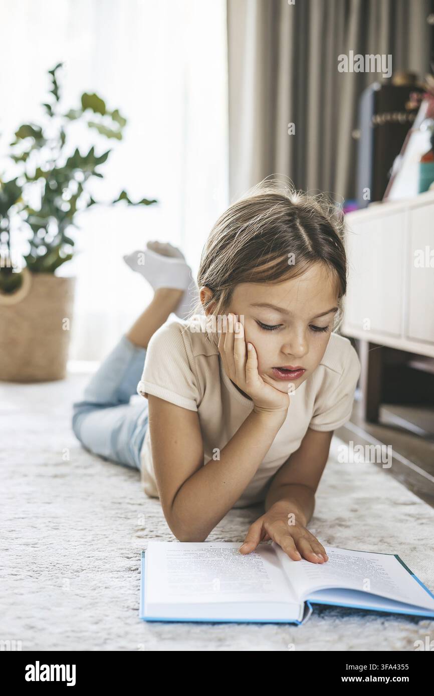Junges Mädchen liegt bequem auf einem gemütlichen Wohnzimmerteppich und liest ein Buch, perfekt für Konzepte des Lernens, der häuslichen Freizeit Stockfoto