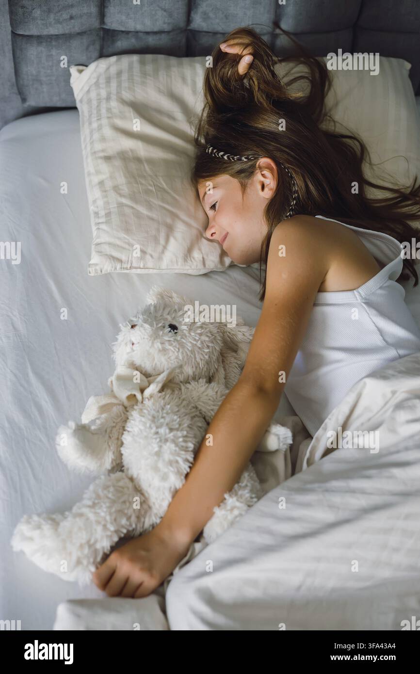 Friedliches junges Mädchen, das in einem gemütlichen Bett schläft und ein großes weißes Spielzeug umarmt, das für die Unschuld der Kindheit steht Stockfoto