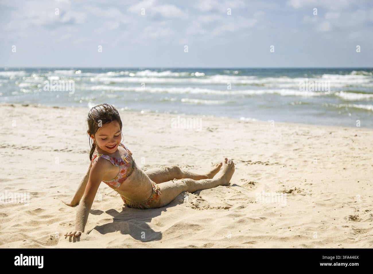 Ein süßes kleines Mädchen im Badeanzug sitzt am sandigen Strand, mit Sand bedeckt, während hinter ihr die Meereswellen brechen ? ein Bild, das die Stockfoto