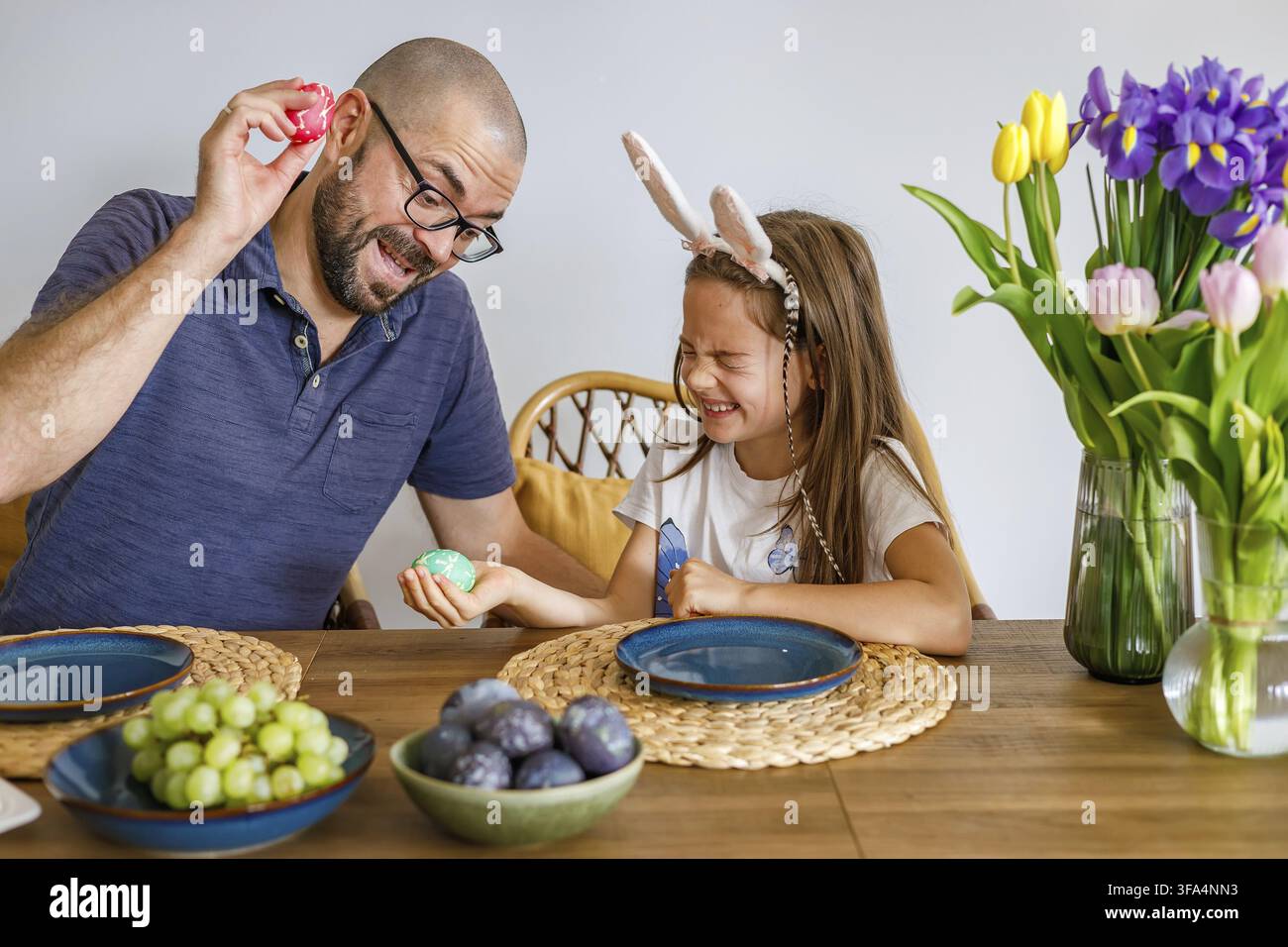 Vater und Tochter spielen ein Klopfspiel mit bunten Ostereiern an einem festlich gedeckten Tisch, der für Familienzusammenhalt und Osterfreude steht Stockfoto
