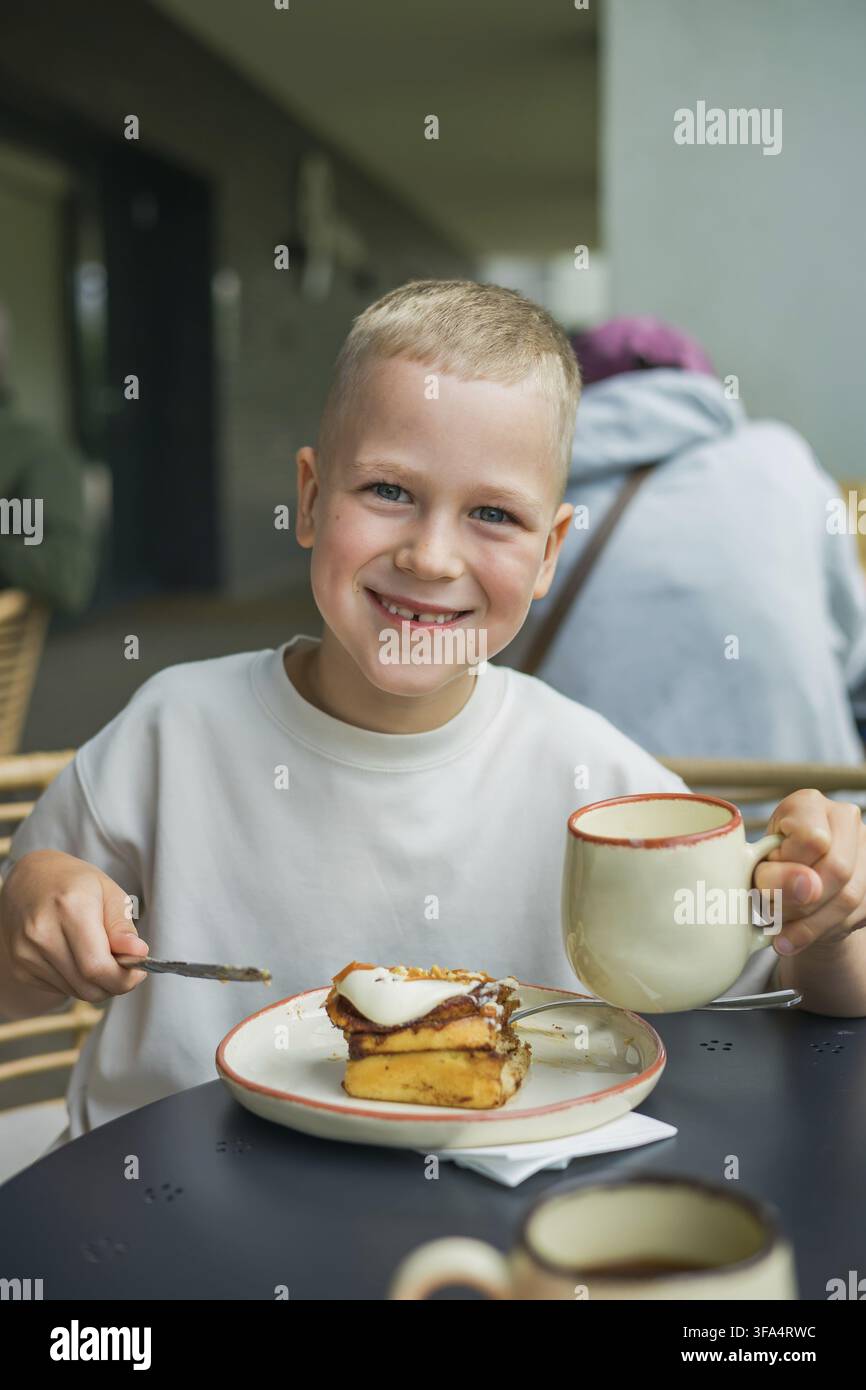 Ein lächelnder Junge, der eine Tasse und ein Messer in der Hand hält, während er in einem Straßencafé ein süßes Brötchen isst Stockfoto