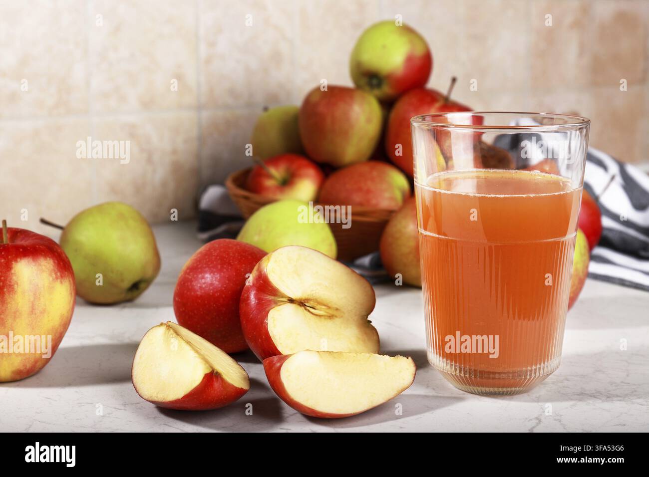 Ein Glas frischer Apfelsaft auf einer Marmorküchenarbeitsplatte, begleitet von ganzen und geschnittenen roten Äpfeln in einem Weidenkorb. Stockfoto