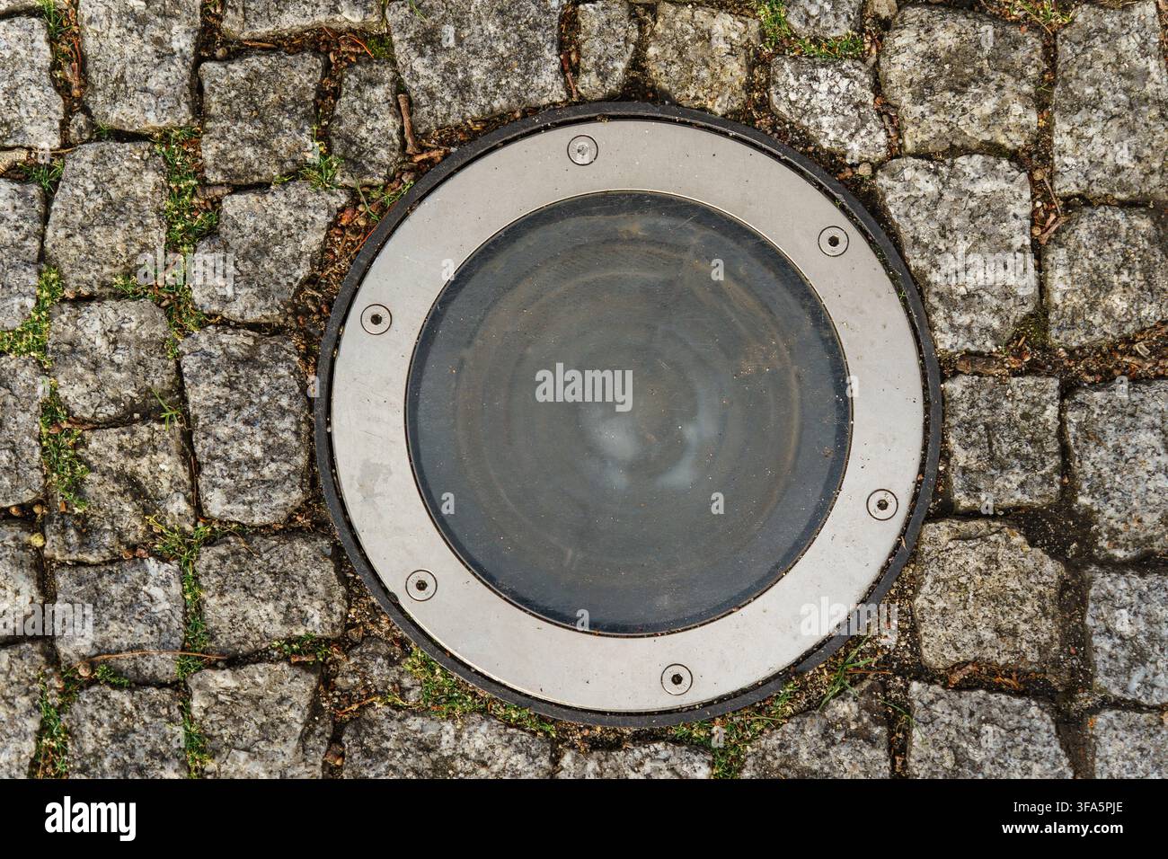 Rundes bodengrabenes Lichtfixture, eingebettet in strukturierten grauen Granitkobelsteinbelag, urbanes Außenbeleuchtungskonzept Stockfoto
