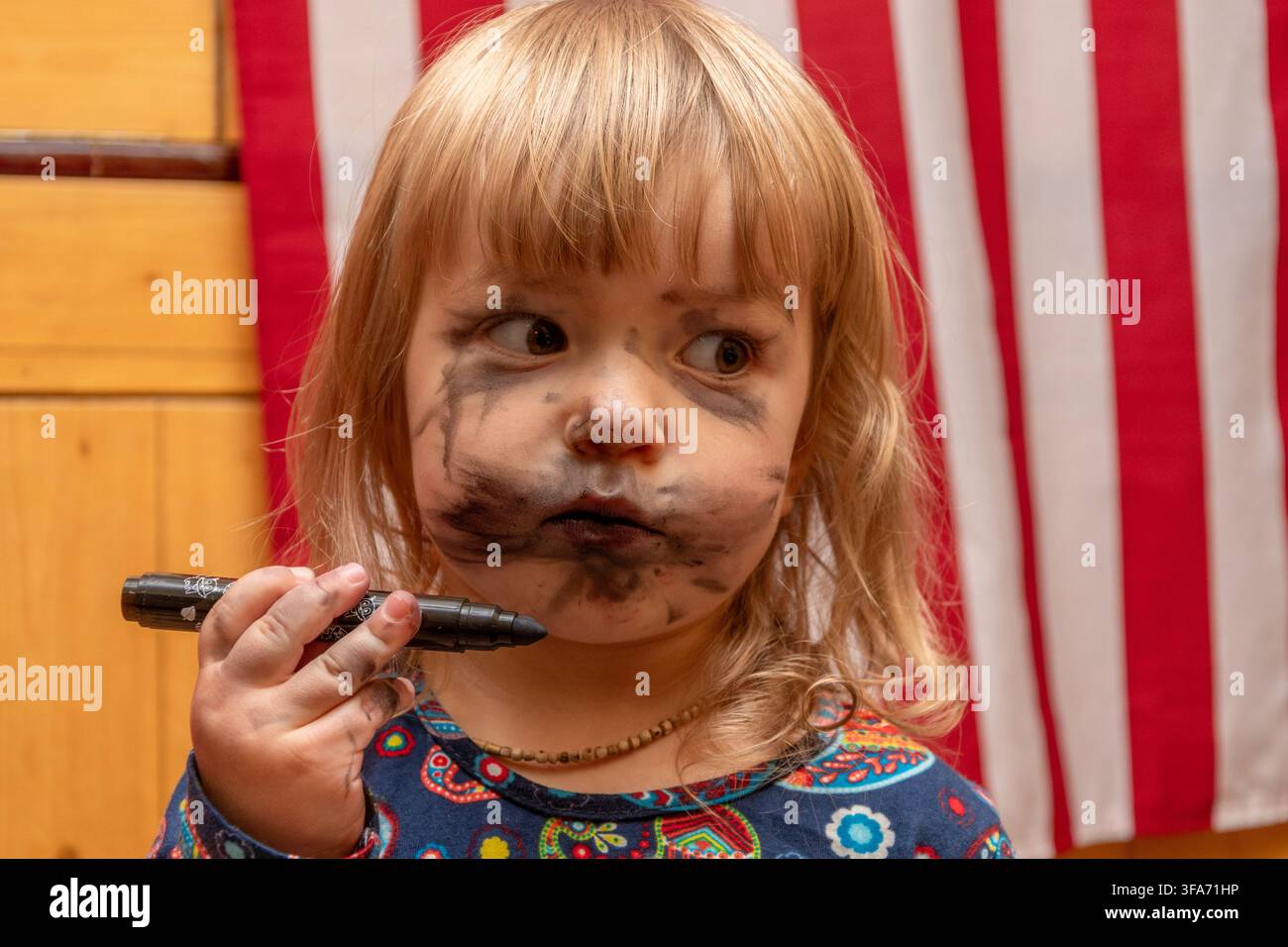 Ein kleines Mädchen mit schwarzem Marker über das Gesicht geschmiert hält den Stift und schaut zur Seite. Stockfoto