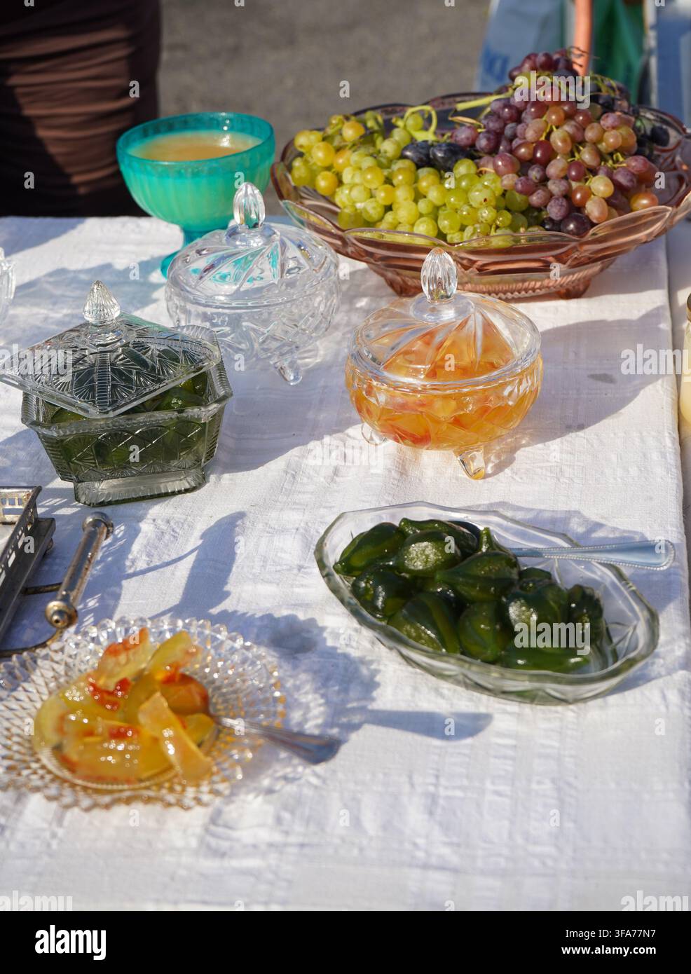 Auswahl an traditionellen griechischen Löffelsüßigkeiten (glyka tou koutaliou) in dekorigem Kristallglas serviert neben frischen Trauben auf einer weißen Tischdecke Stockfoto