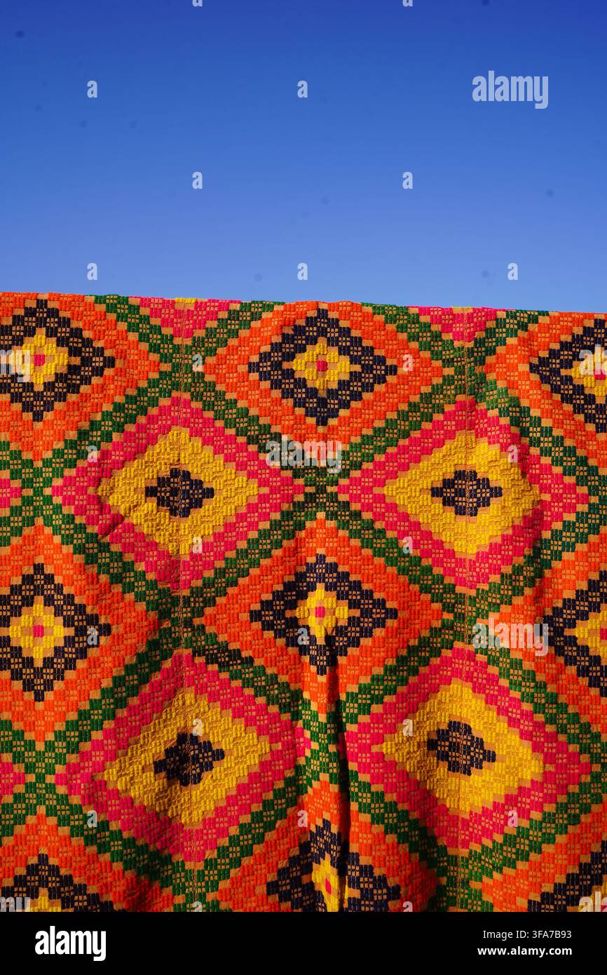 Vertikale Aufnahme eines lebendigen handgewebten Kilim-Teppichs mit bunten Diamantmustern, der im Freien unter einem klaren blauen Himmel hängt. Traditionelle griechische Volkskunst. Stockfoto