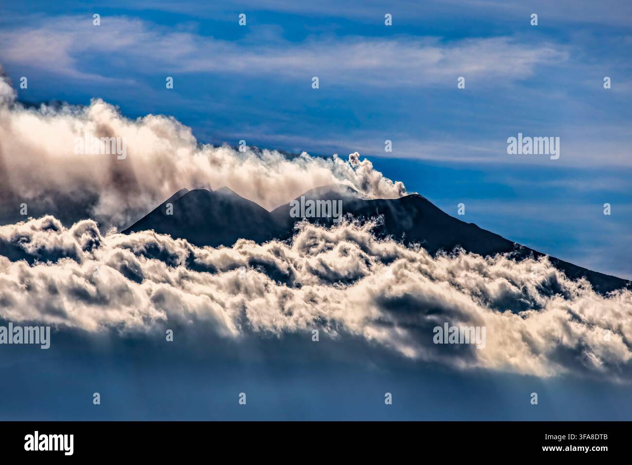 Der Ätna erhebt sich über einem Meer von Wolken. Der dunkle Vulkanberg steigt dramatisch durch wallende weiße Wolken unter einem lebendigen blauen Himmel. Stockfoto