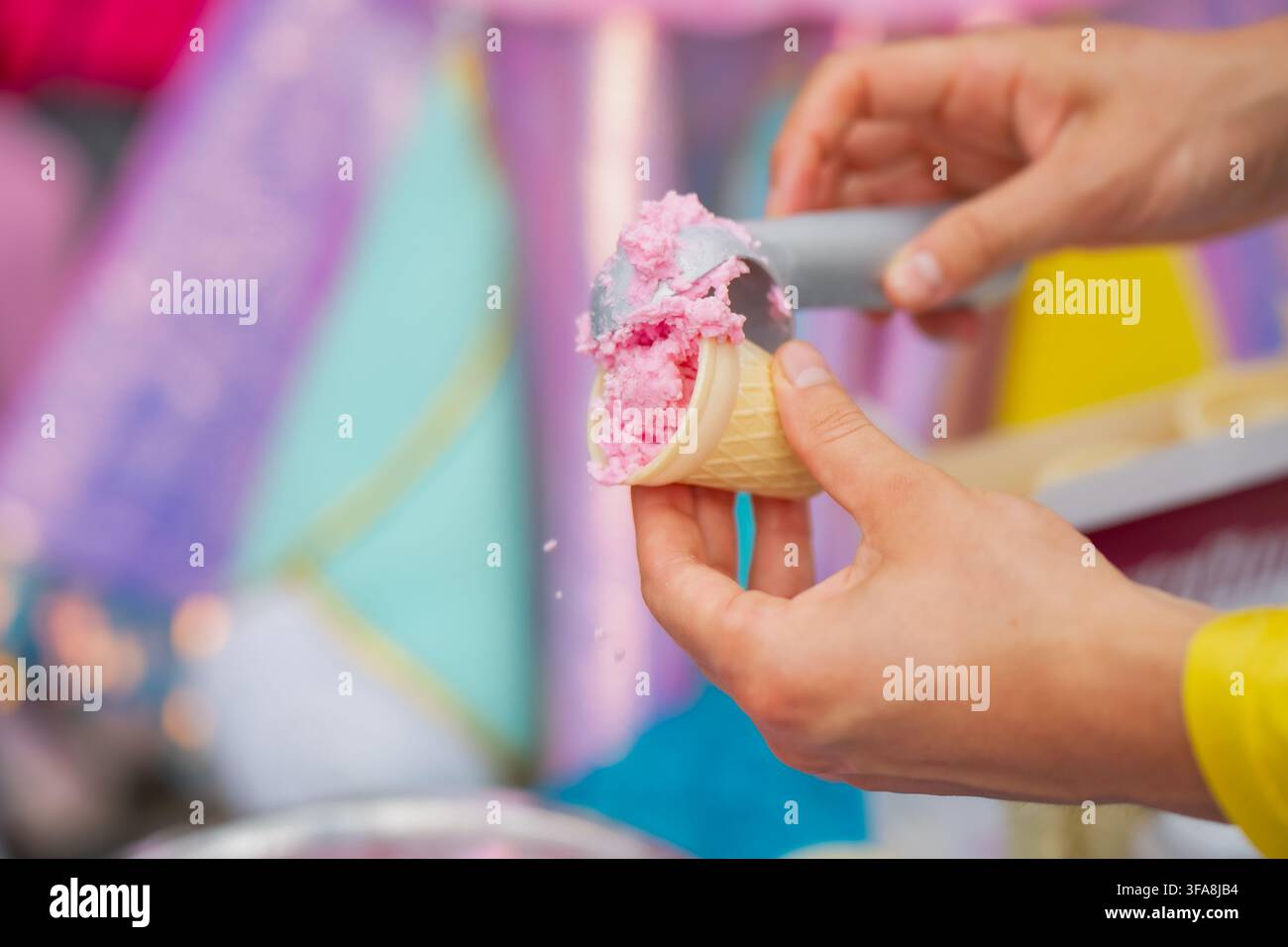 Person serviert cremiges rosa Fruchtgelato-Eis in eine kleine Waffelkegel mit einem Metall-Eislöffel für ein sommerliches Erfrischungs-Event Stockfoto