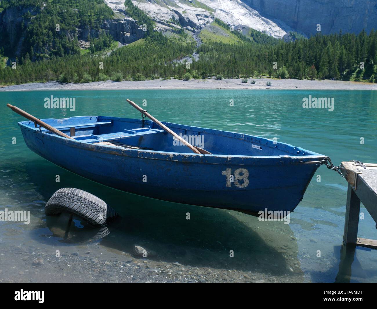 Ein blaues Holzruderboot ruht am Ufer eines türkisfarbenen Alpenssees, umgeben von Bergen, bereit zur Vermietung. Perfekt für Reisen, Freizeit und die Schweiz Stockfoto