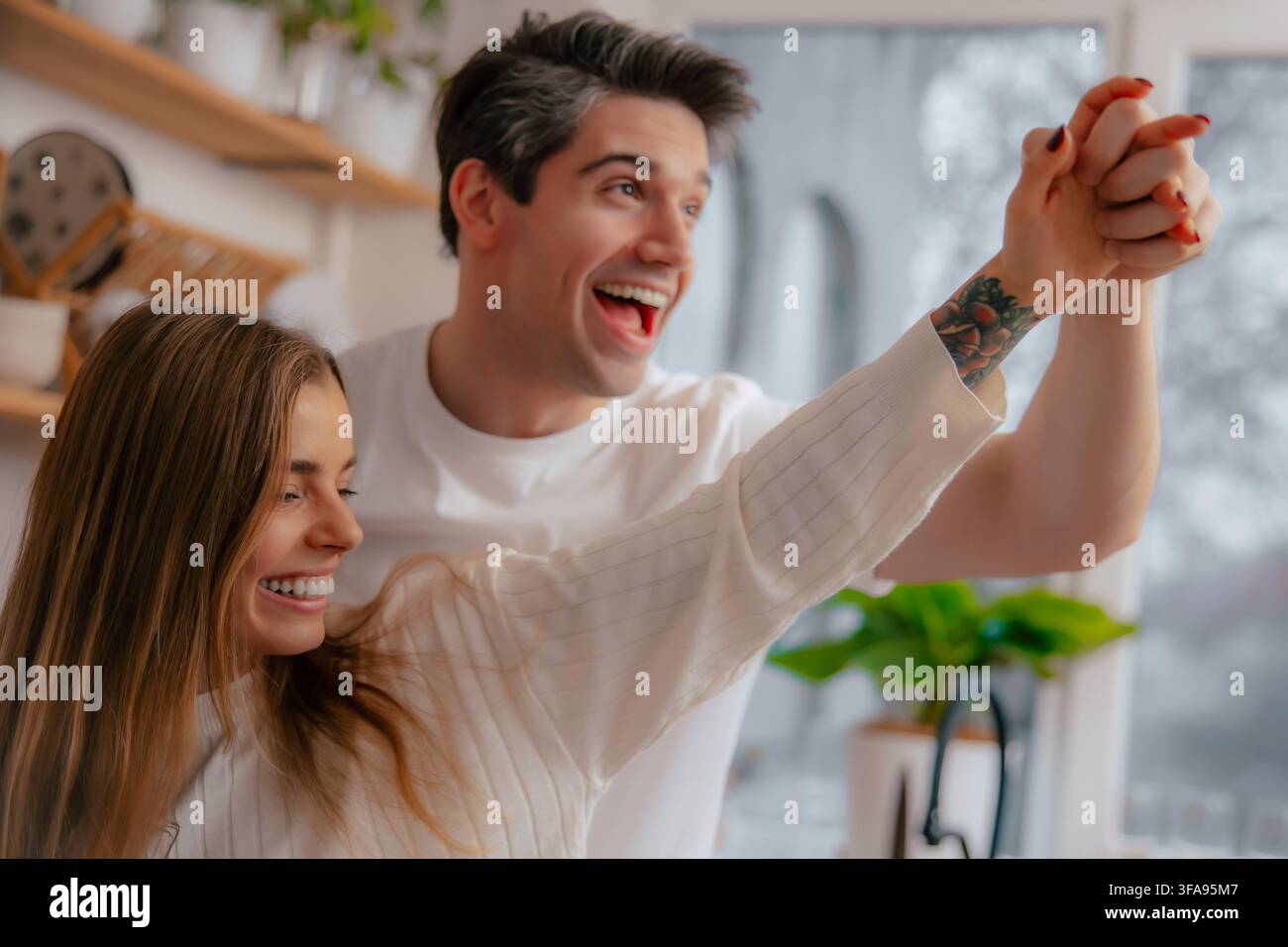 Nahaufnahme eines fröhlichen Paares, das seine verschränkten Hände erhebt, während es zusammen in einer hellen Küche lacht und tanzt. Stockfoto