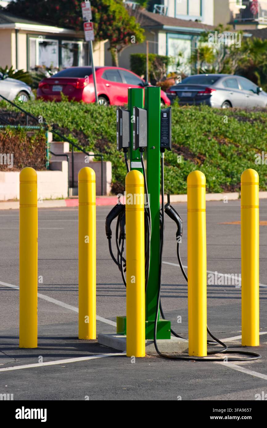Grüne Elektroauto-Ladestation steht auf einem Parkplatz. Gelbe Poller umgeben die Station zur Sicherheit. Autos parken in der Nähe in einer sonnigen Außenumgebung Stockfoto