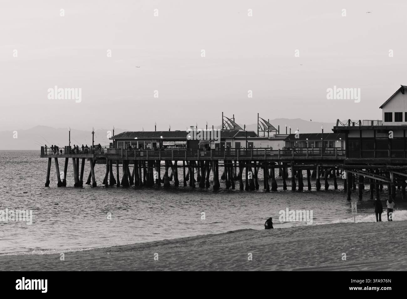 Schwarz-weißer Pier erstreckt sich über ruhiges Meer. Menschen gehen am Ufer und Pier entlang. Die Atmosphäre wirkt friedlich und nachdenklich. Gut für Reisen, Küsten-, Stockfoto