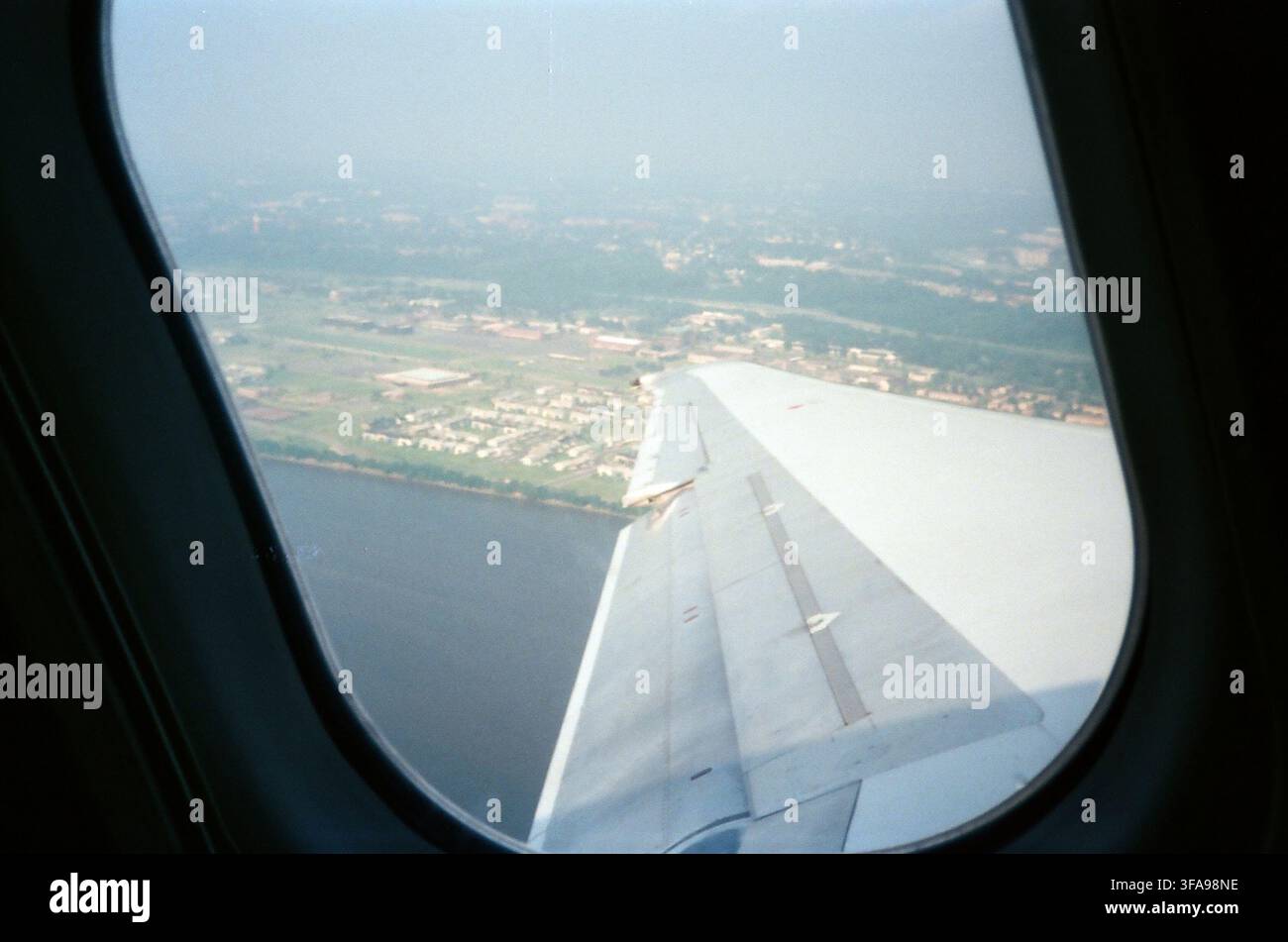 Blick aus dem Fensterplatz des Flugzeugs, der den Flugzeugflügel mit ausgefahrenen Klappen über einem Fluss und einer grünen städtischen Landschaft während des Anfluges oder Starts zeigt Stockfoto