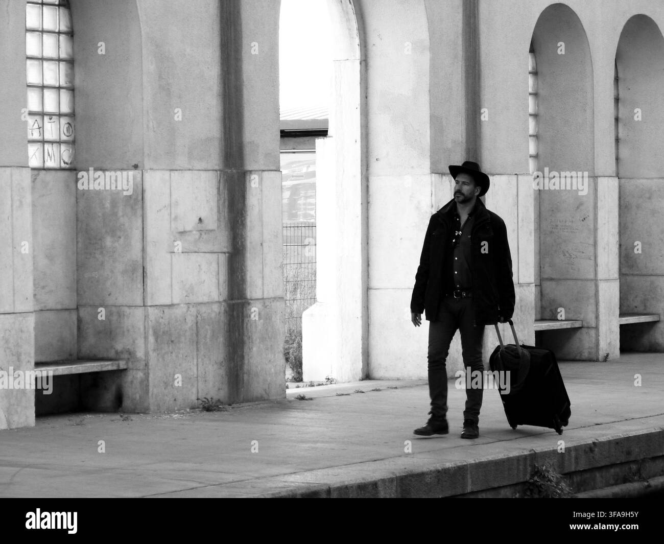 Ein stilvoller, dunkelhaariger, schlanker, bärtiger weißer Mann von 40 Jahren, der einen Cowboyhut trägt, bewegt sich mit einem Rollkoffer Stockfoto