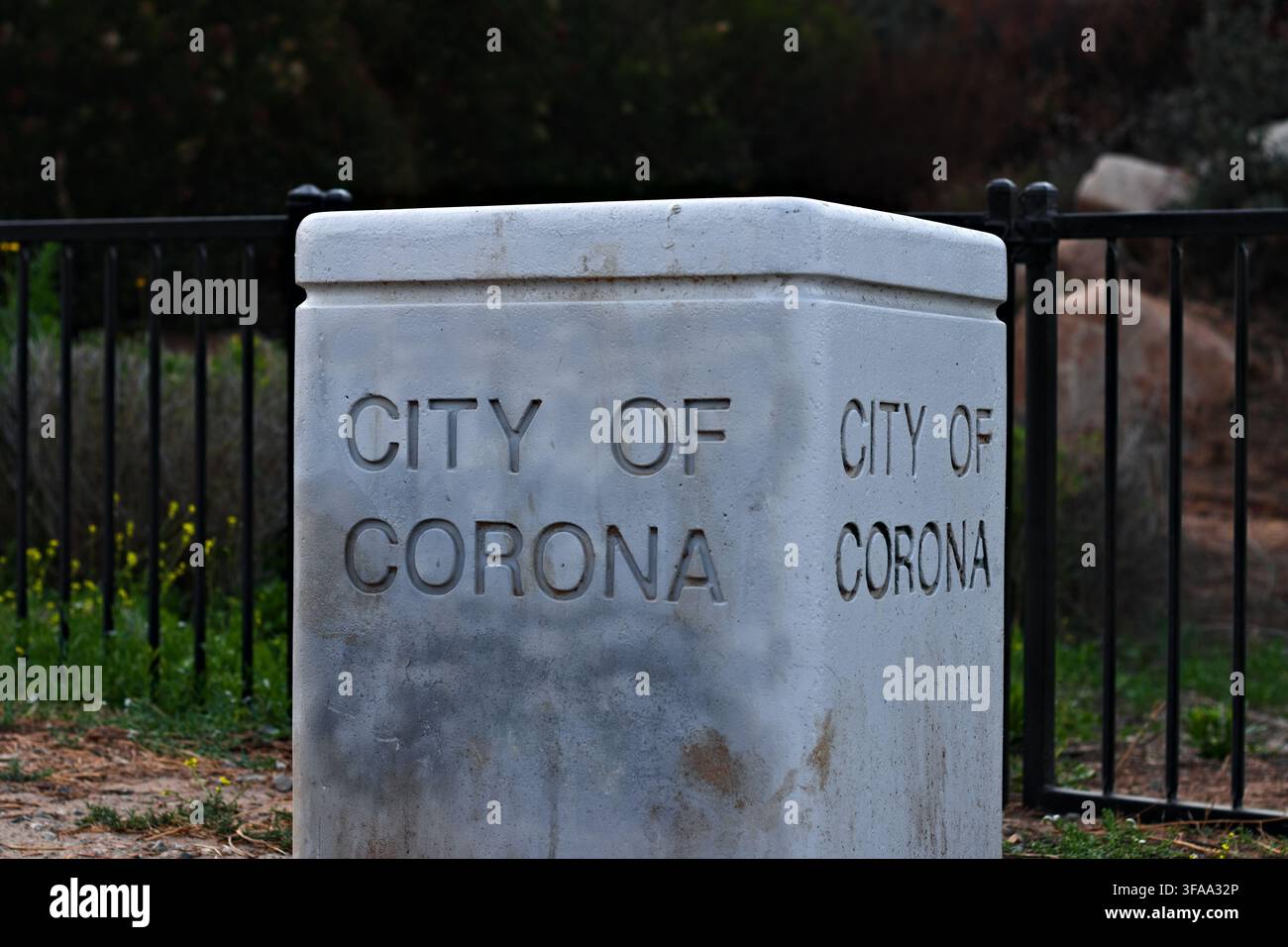 Steinerne Markierung der Stadt Corona steht in der Nähe eines schwarzen Metallzauns. Verwitterter Beton trägt den eingravierten Stadtnamen in klaren Buchstaben. Grünes Gras und Wildblumen s Stockfoto