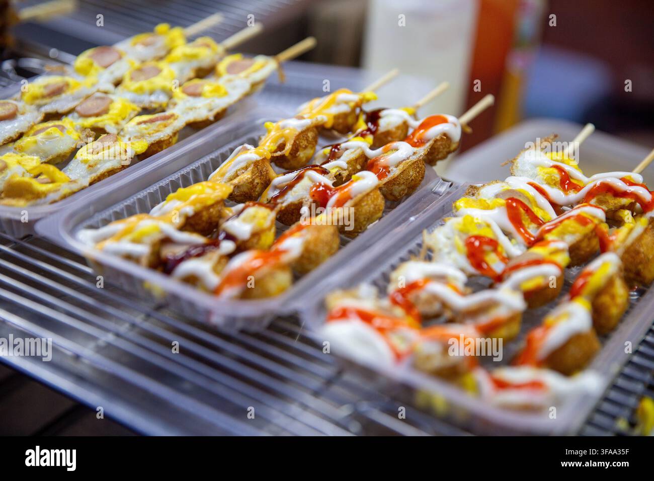 Reihen von tiefgebratenen Straßenimbiss-Spießen und gebratenen Wachteleierstäbchen, beträufelt mit Mayonnaise und Chilisauce, die in Plastikbehältern im Freien stehen Stockfoto