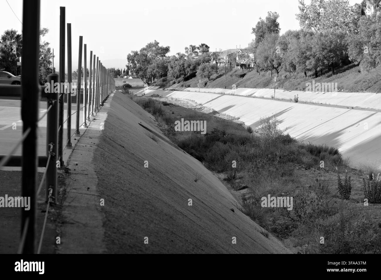 Ein trockener Beton-Überwasserkanal verläuft neben einem Parkplatz und einem landschaftlich gestalteten Hügel, gesäumt von Barrieren, Unkraut, Bäumen und nahe gelegenen Häusern. Stockfoto