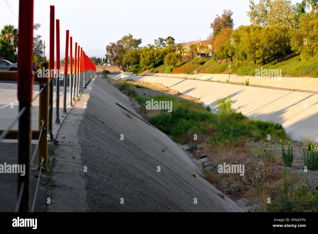 Ein trockener Beton-Überwasserkanal verläuft neben einem Parkplatz und einem landschaftlich gestalteten Hügel, gesäumt von Barrieren, Unkraut, Bäumen und nahe gelegenen Häusern. Stockfoto