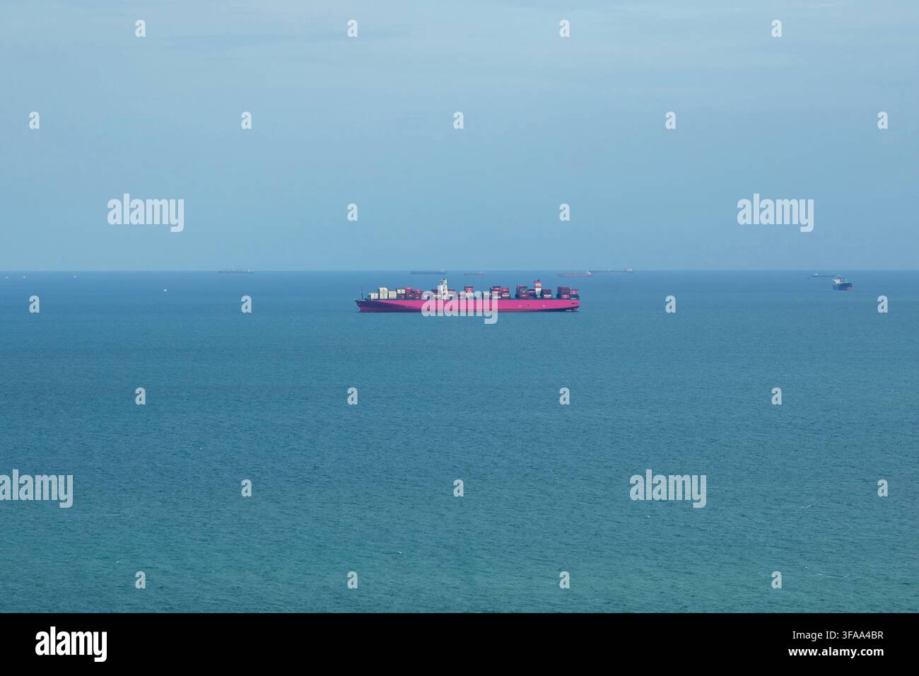 Rosa Containerschiff fährt auf ruhiger blauer See nahe der Küste unter einem dunstigen Himmel, Konzept für Seeschifffahrt und Transport Stockfoto