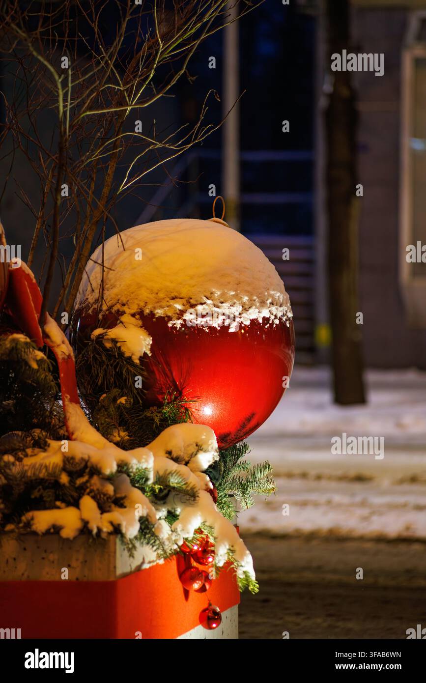 Ein riesiger glänzender roter Weihnachtskugel-Ornamente steht auf immergrünen Kiefernzweigen, die mit frischem Schnee bedeckt sind. Die festliche Straßendekoration wird von warmem Licht beleuchtet Stockfoto