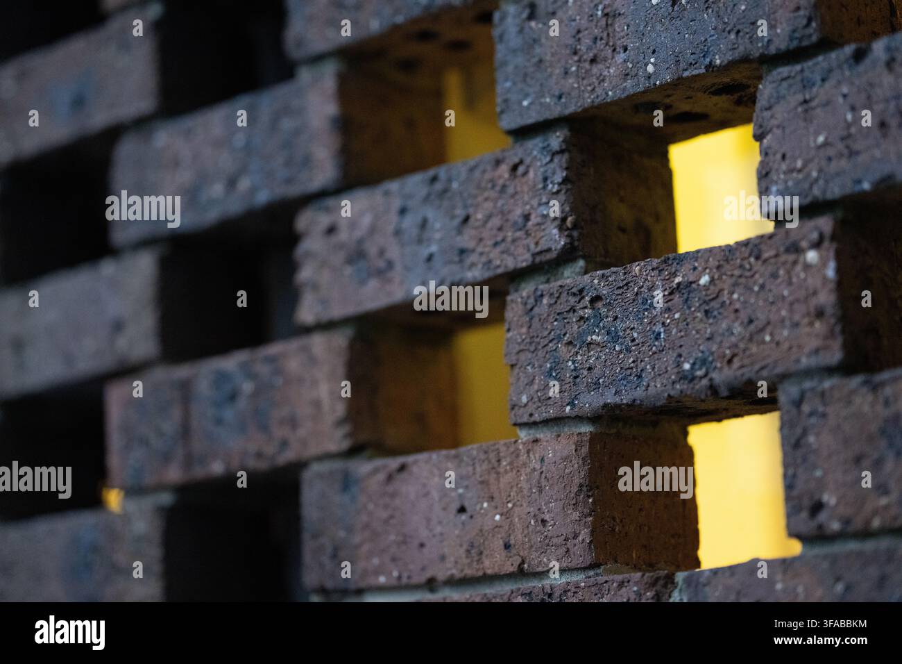 Nahaufnahme einer dunklen Ziegelsteinwand mit warmem gelbem Licht, das durch Lücken scheint, Architektur- und Designdetail mit flacher Tiefenschärfe Stockfoto