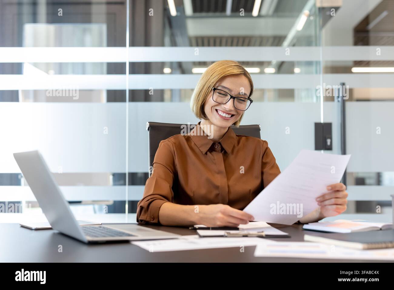 Zufriedene Geschäftsfrau liest Unterlagen an einem Bürotisch, lächelt, während sie Dokumente hält und an ihrem Laptop arbeitet, zeigt Arbeitszufriedenheit und Erfolg in ihrer Karriere Stockfoto