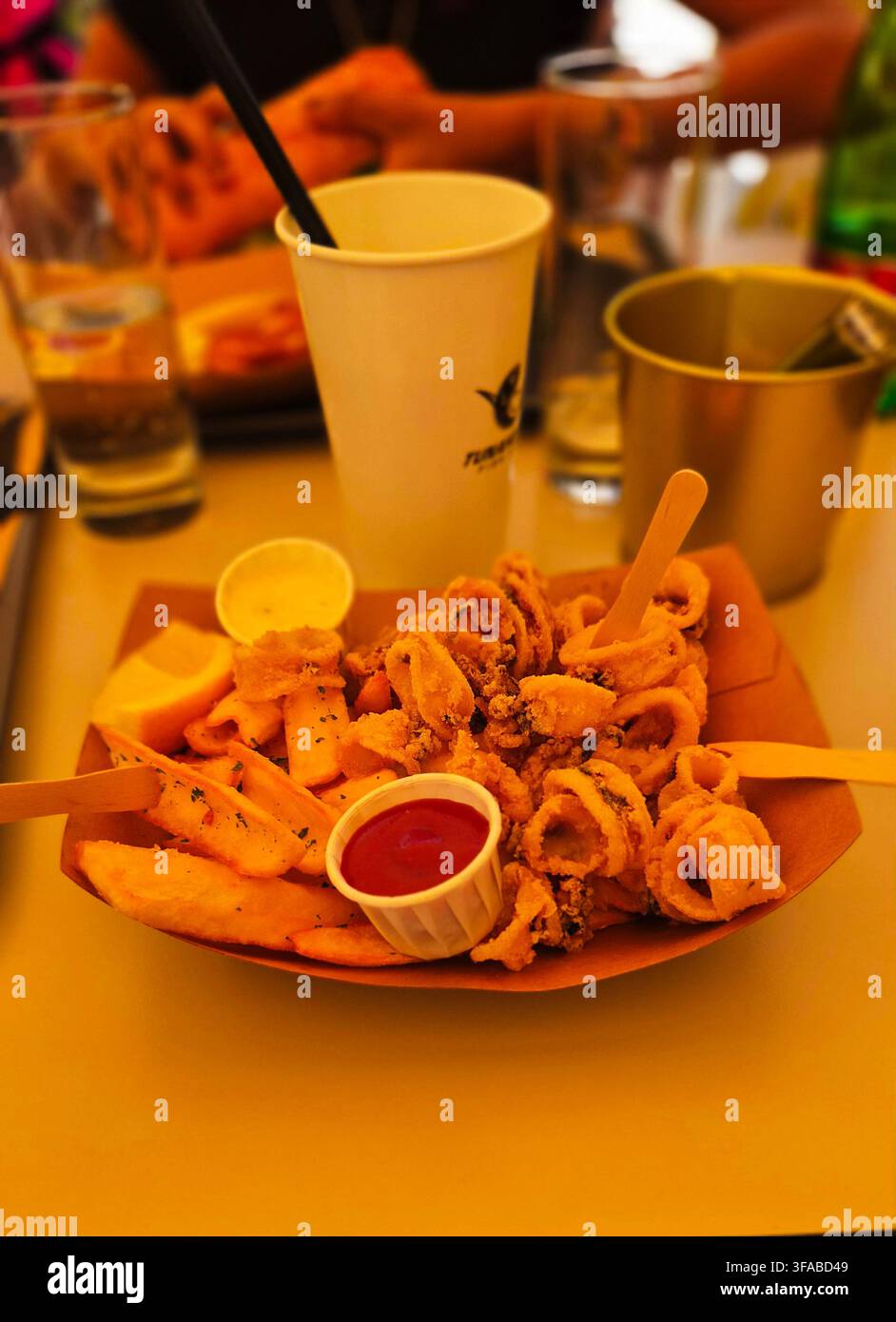 Beschriftungen Redaktionell / Beschreibend: Knusprig gebratene Calamariringe und Kartoffelkeile, serviert in einem Kartontray mit Dipsaucen, Zitronenspalter und Getränk Stockfoto