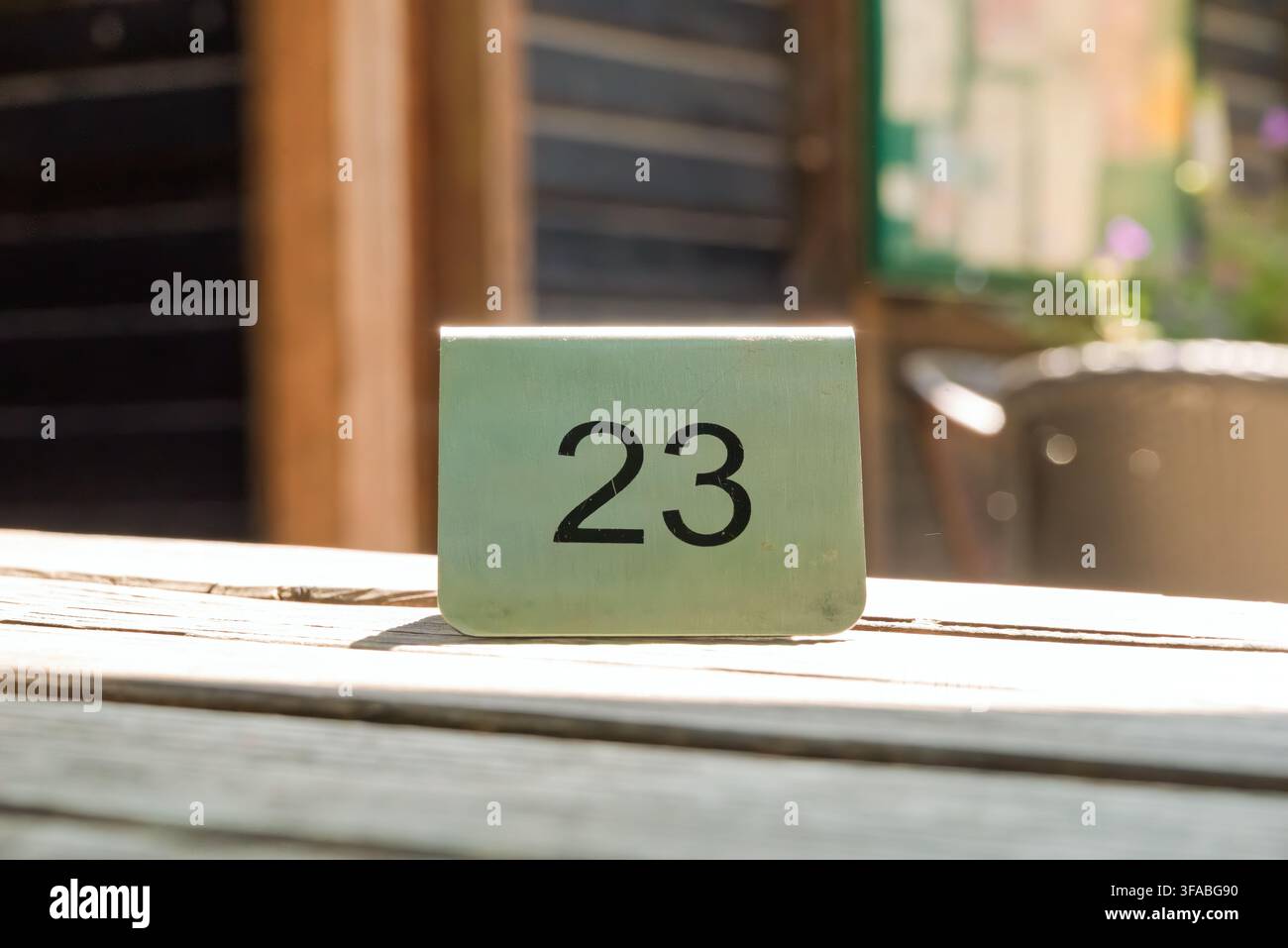 Grüne Tischnummer 23 auf einer Holzoberfläche. Sonnenlicht erzeugt Highlights. Unscharfer Hintergrund zeigt Gebäude und Grün. Ort: Außenbereich eines Cafés Stockfoto