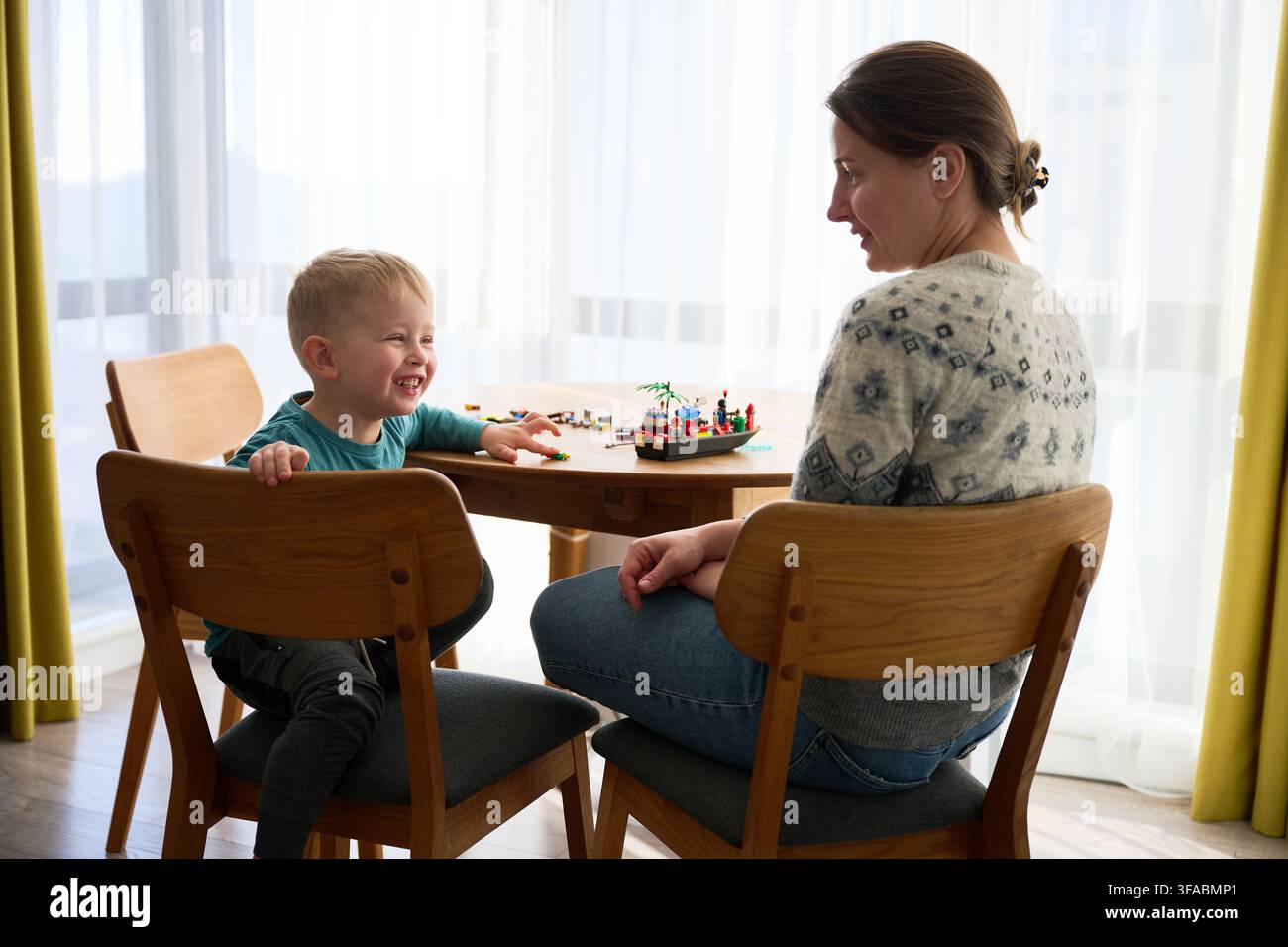 Junge spricht und lacht mit der Mutter. Sitzt zu Hause am Tisch, spielt mit Bauklötzen, warme Familieninteraktion und glücklicher Moment der Kindheit Stockfoto