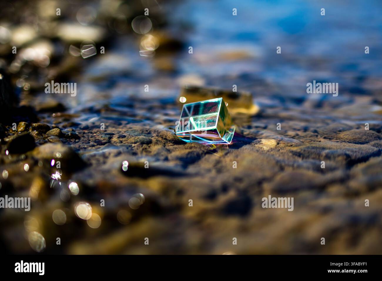 Kleines Glasprisma, das auf flachen Flusssteinen ruht, fängt Sonnenlicht ein und wirft helle Spektralfarben, während sanfte Wellen um es herum mit träumerischem Glitzern funkeln Stockfoto