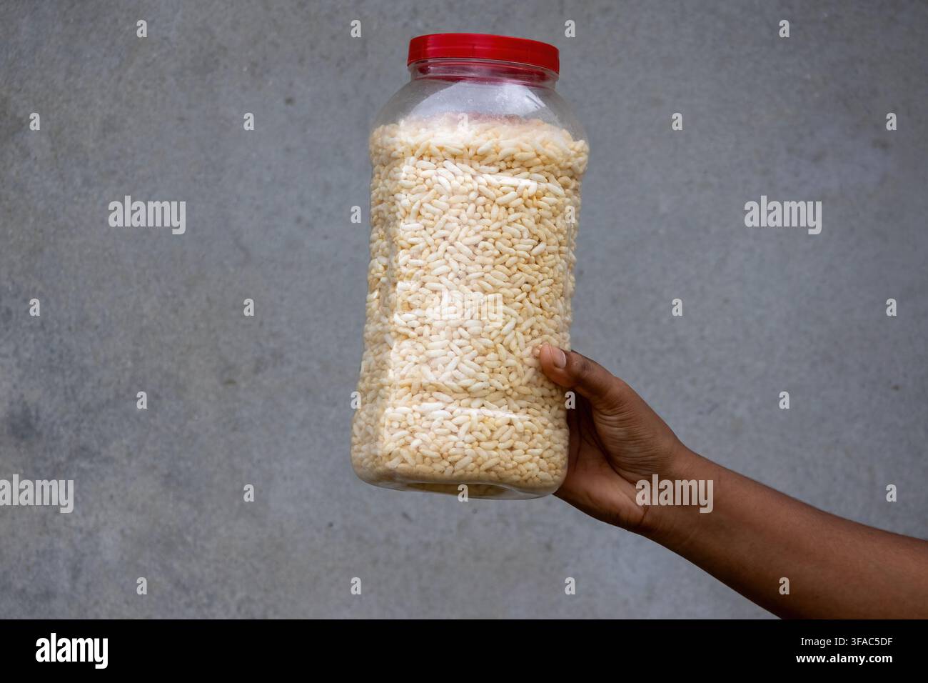 Person hält einen Plastikbehälter gefüllt mit knusprigem Puffreis vor rustikalem grauem Wandhintergrund. Traditionelle südasiatische Muri, die für den täglichen Tee aufbewahrt wird Stockfoto