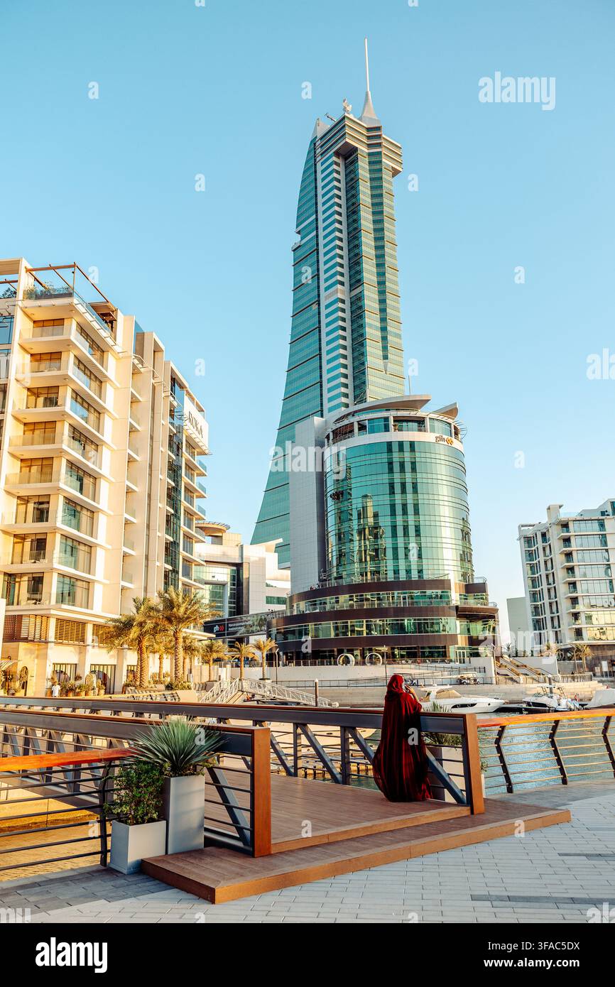 Manama, Bahrain - 13. Juni 2026: Blick auf die moderne Skyline mit den Bahrain Financial Harbour-Türmen, mit einer Frau in einem Abaya, die auf einem Fuß steht Stockfoto