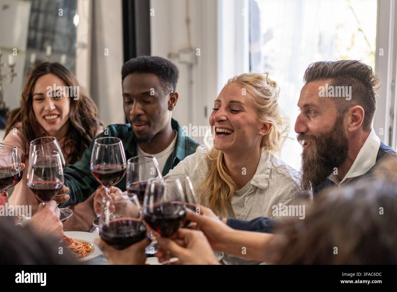 Multikulturelle Freunde stoßen mit Gläsern roten Weins an und lachen gemeinsam während eines intimen Abendessens zu Hause. Gen Z und Millennials teilen eine Stockfoto