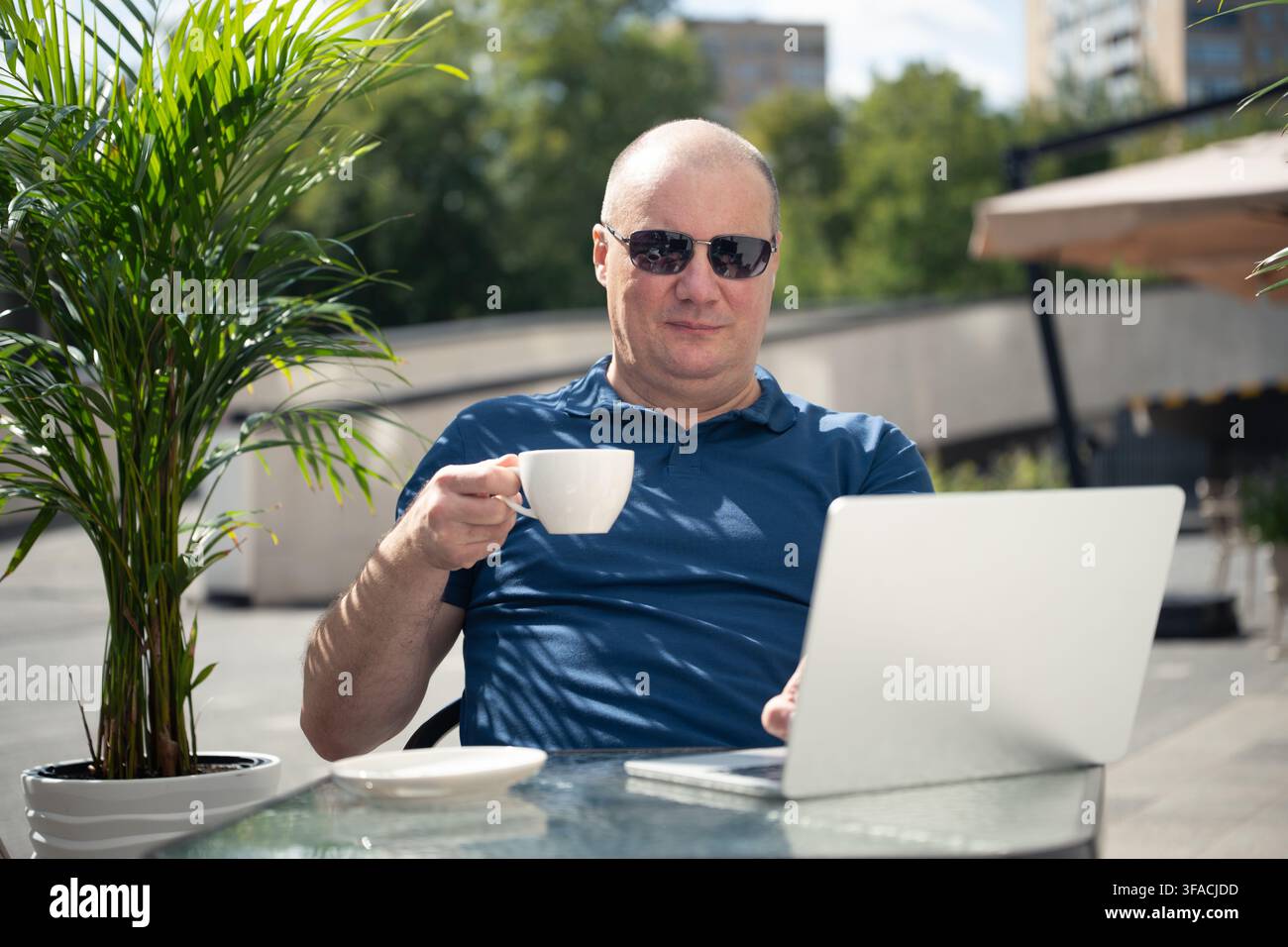 Reifer kaukasischer Mann verwendet einen Laptop mit Dokumenten, Stift und Kaffee auf dem Tisch, während er in einem Outdoor-Café remote arbeitet. Stockfoto