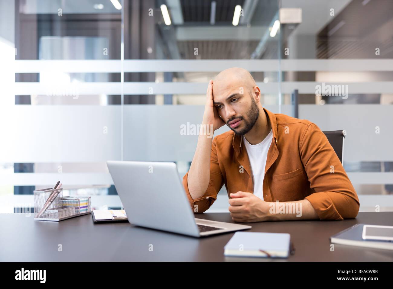 Junger Mann sitzt an seinem Schreibtisch und schaut mit einem gestressten und überforderten Ausdruck auf seinen Laptop-Bildschirm, während er über seine Arbeit nachdenkt. Stockfoto