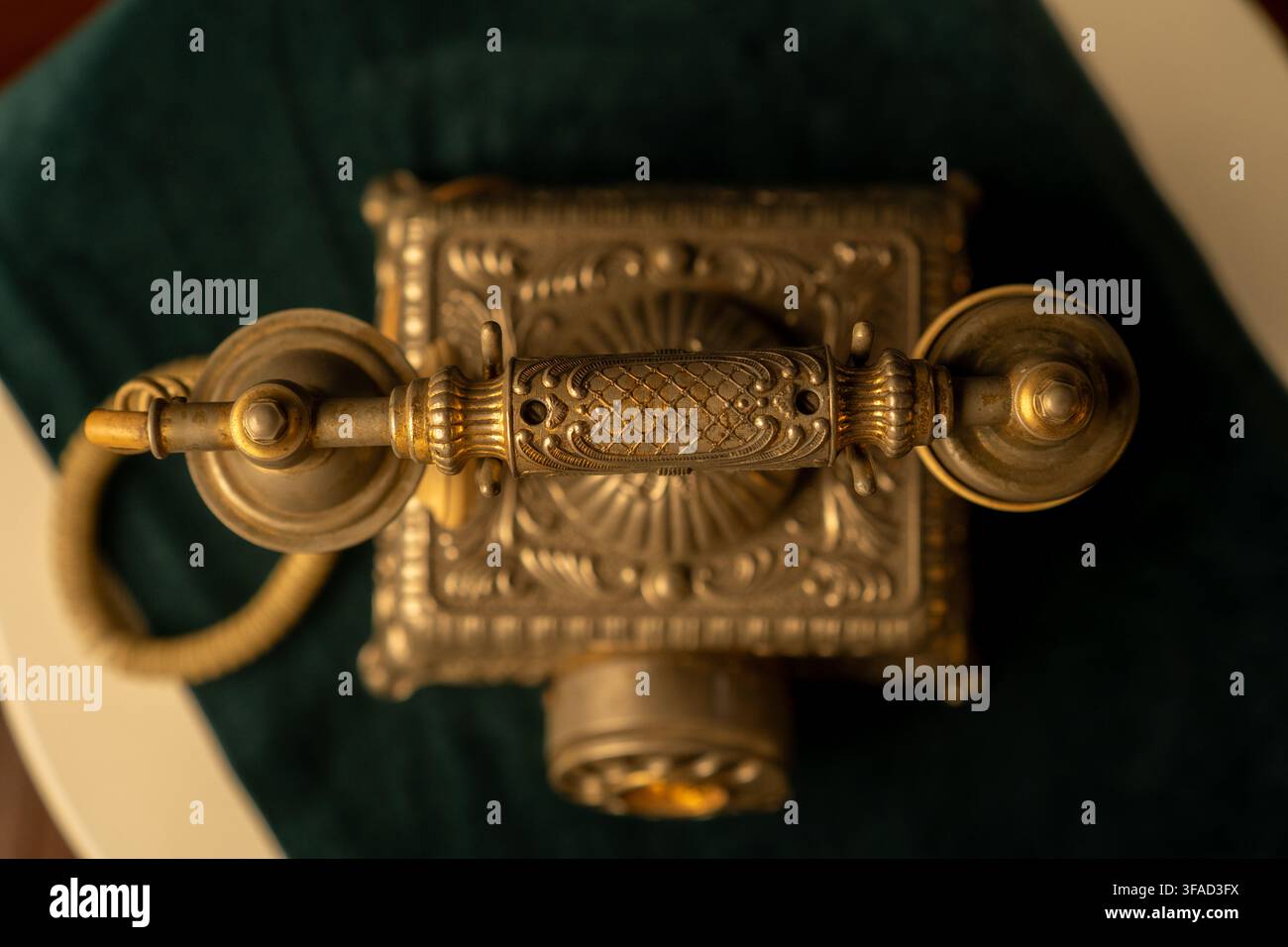 Aufsicht von oben auf ein altes, verziertes Messing-Telefonhörer, das auf seiner Basis ruht, mit symmetrischen Metallmustern, warmen Highlights und dunklen Stockfoto