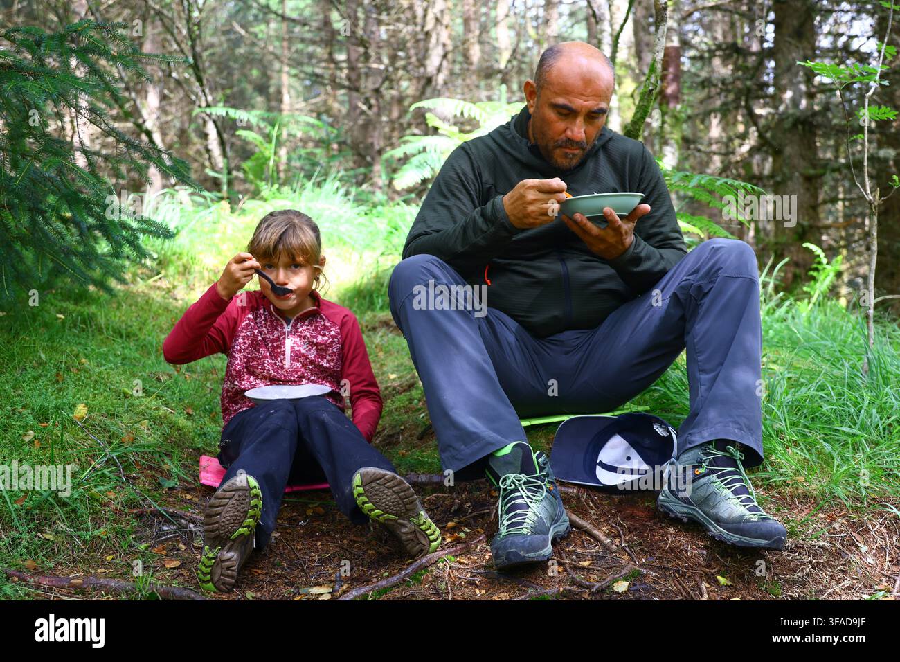 Vater und Kind sitzen auf weichem Waldboden und essen warme Speisen aus Schüsseln, umgeben von Farnen und hoher Vegetation. Der gemeinsame Moment spiegelt das E Stockfoto
