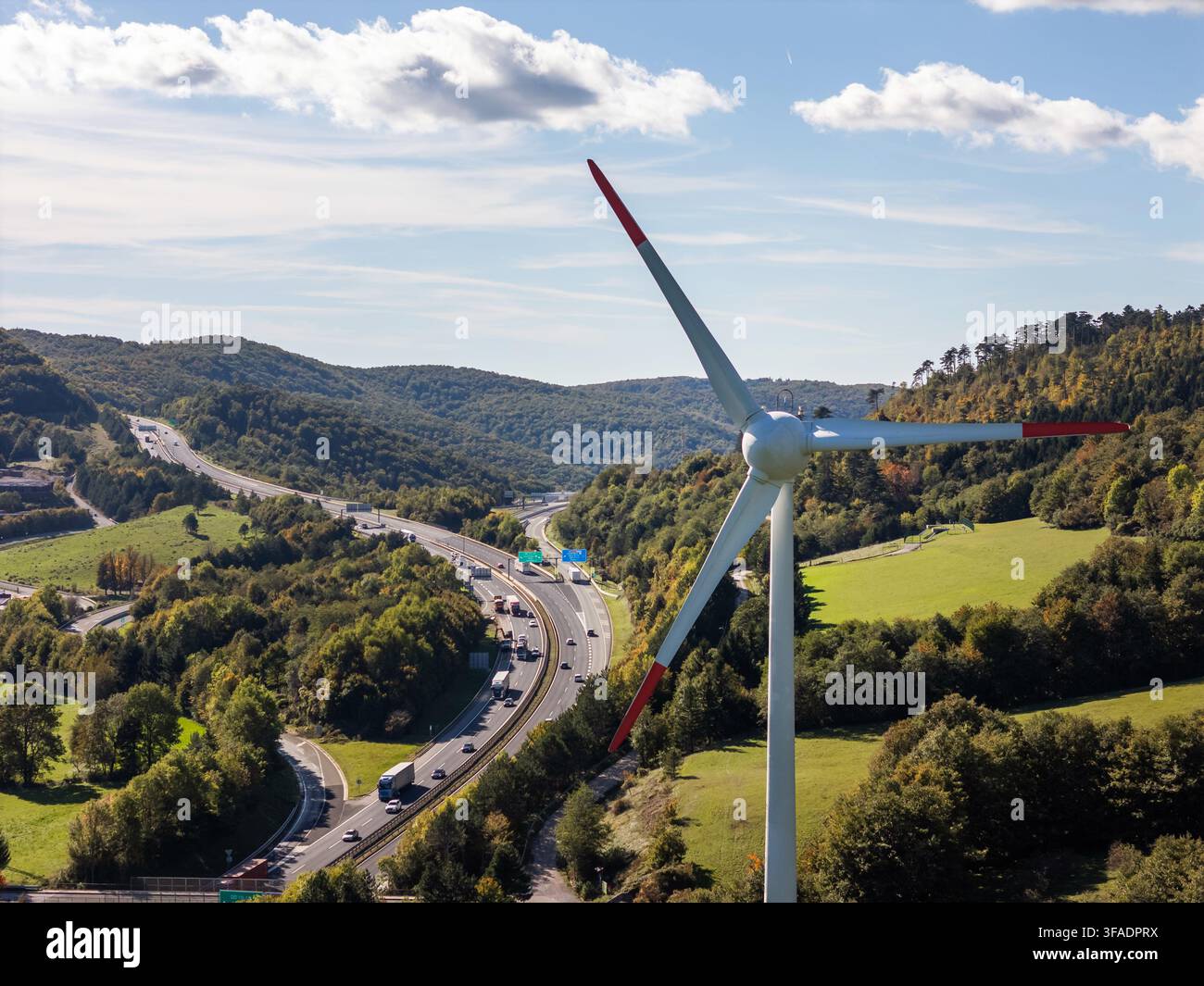 Windturbine steht hoch neben einer belebten Autobahn mit Fahrzeugen. Grüne Hügel füllen den Hintergrund an einem sonnigen Tag. Stockfoto
