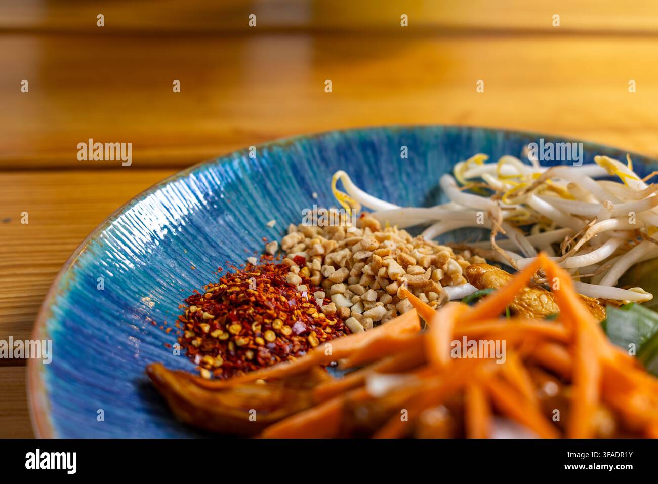 Zutaten der asiatischen Küche liegen auf einem blauen Keramikteller, mit Chiliflocken, gehackten Erdnüssen, Mungobohnensprossen, Karottenstiften und einem gekochten Gemüse Stockfoto