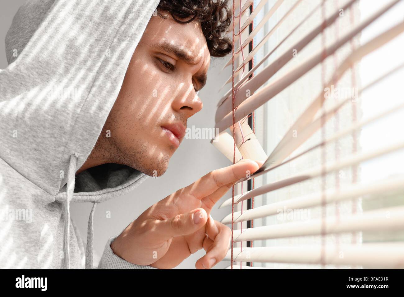 Datenschutz- und Überwachungskonzept, junger Mann in Kapuzenpullover späht durch Fensterläden, fühlt sich beobachtet oder misstrauisch Stockfoto